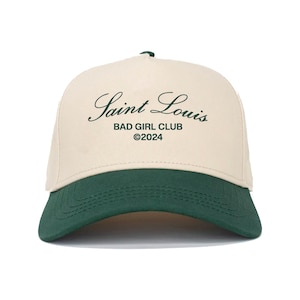 Puede incluir: Una gorra de béisbol beige y verde con el texto "Saint Louis Bad Girl Club ©2024" bordado en la parte delantera.