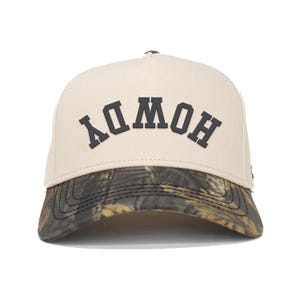 May include: A beige baseball cap with the word "HOWDY" in dark grey block letters. The brim has a camouflage pattern in shades of brown, green, and black. The cap is a classic six-panel design.