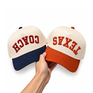 May include: Two baseball caps with off-white crowns. The left cap features a navy blue brim and the word COACH in red embroidery. The right cap features an orange brim and the word TEXAS in orange embroidery. Both caps have a small button on top.