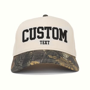 Könnte beinhalten: Beige und Camouflage Baseballkappe. Auf der Vorderseite der Kappe steht in fetten schwarzen Buchstaben das Wort "CUSTOM", darunter "TEXT". Der Schirm hat ein Camouflagemuster.