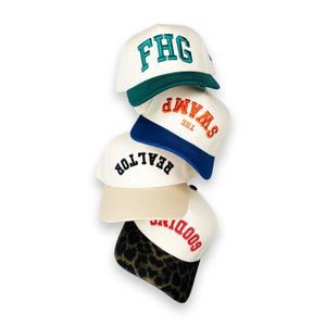 Puede incluir: Una pila de cuatro gorras de béisbol con coronas blancas. La gorra superior tiene una visera verde oscuro y el texto FHG en color verde azulado. La segunda tiene una visera azul y el texto THE SWAMP en naranja. La tercera tiene una visera beige y el texto REALTOR en negro. La inferior tiene una visera con estampado de leopardo y el texto GOODTIMES en rojo.