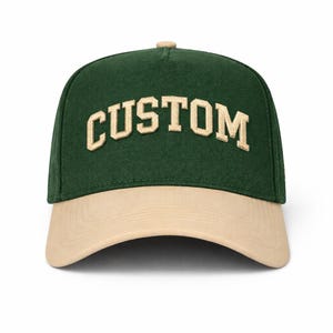 Pode incluir: Boné de beisebol verde e bege com a palavra "CUSTOM" bordada em letras de estilo bloco bege na frente. O boné tem uma viseira curva e um botão na parte superior.
