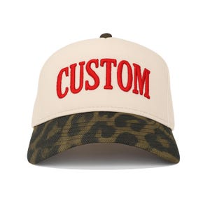 May include: A cream-coloured baseball cap with the word "CUSTOM" embroidered in red across the front. The brim features a brown and olive green animal print. The cap has a dark brown button on top.