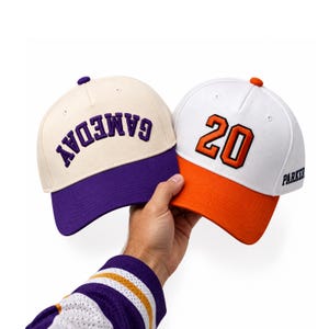 Puede incluir: Dos gorras de béisbol. La gorra izquierda es de color crema con visera morada y el texto GAMEDAY bordado en morado. La gorra derecha es blanca con visera naranja, el número 20 bordado en naranja y el texto PARKER en el panel lateral.