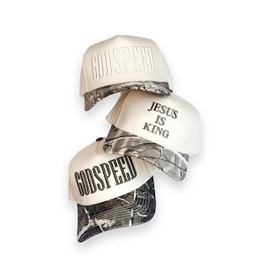 May include: A stack of cream-coloured baseball caps with camouflage brims. The caps feature the embroidered text "GODSPEED" and "JESUS IS KING" in black. The caps are stacked on a white background.