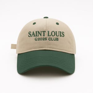 May include: A beige and green baseball cap with the text "SAINT LOUIS C2025 CLUB" embroidered on the front. The cap has a curved brim and an adjustable strap on the back.