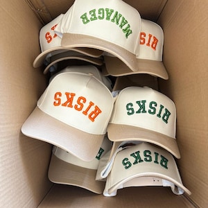 May include: A box full of beige baseball caps. The caps have the word "BISKS" embroidered in green and orange on the front. One cap has the word "MANAGER" embroidered in green on the front.
