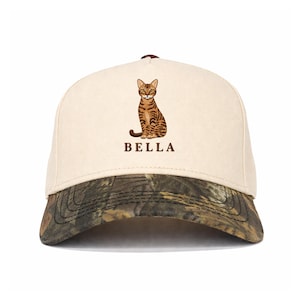 May include: A baseball cap with a cream-coloured front panel and a dark camouflage-patterned brim. The front features a brown tabby cat illustration centred above the text BELLA in brown capital letters. The top button is a dark brown colour.