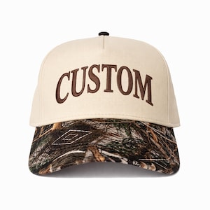 Puede incluir: Gorra de béisbol beige con la palabra "CUSTOM" bordada en marrón. La visera presenta un estampado de camuflaje en tonos marrones, verdes y beige. La gorra tiene un botón marrón oscuro en la parte superior.