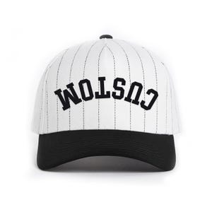 Puede incluir: Gorra de béisbol blanca con finas rayas negras y visera negra. La palabra "CUSTOM" está arqueada en la parte delantera en letras negras en negrita. La gorra tiene una visera curva y un botón en la parte superior.