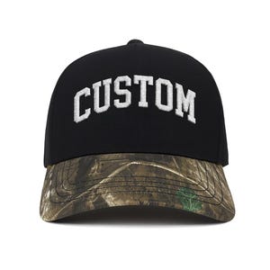 May include: Black baseball cap with the word "CUSTOM" embroidered in white block letters. The brim features a camouflage pattern in shades of brown, green, and tan. The cap has a curved brim and a button on top.