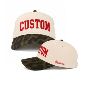 May include: A cream-coloured baseball cap featuring the word CUSTOM in bold red embroidered letters on the front panel. The brim displays a dark brown and black leopard print pattern. A smaller red script version of the word Custom is on the side.