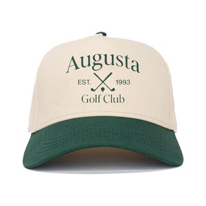 Può includere: Cappellino da baseball beige e verde con la scritta "Augusta Golf Club" in verde. Il cappellino ha una visiera curva e un pannello frontale strutturato. Il testo include l'anno "1993" e mazze da golf incrociate.
