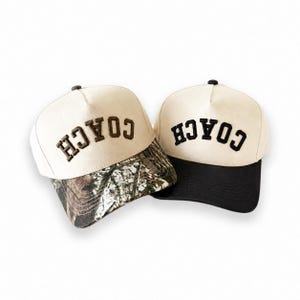 May include: Two baseball caps with off-white crowns and the word COACH embroidered in dark lettering. One cap features a camouflage print on the brim, while the other cap has a solid black brim. Both caps have a structured front panel design.