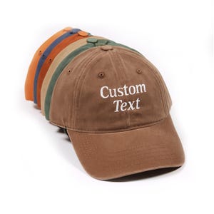 Peut inclure: Plusieurs casquettes de baseball de couleurs variées, dont marron, vert, orange et bleu. La casquette marron au premier plan porte l'inscription "Custom Text" brodée en blanc. Les casquettes sont en tissu doux.