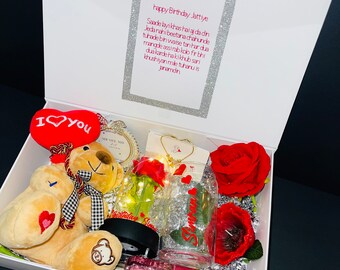 May include: A white gift box filled with various items. Includes a plush teddy bear, a red heart-shaped pillow with "I love you" text, a glass with text, a necklace, a rose, and a bottle of champagne toast.
