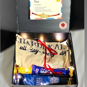 以下が含まれることがあります： Tシャツ、Cadbury Dairy Milkチョコレートバー、Deep Oceanボディクリームが入ったギフトボックス。 「Veer」という言葉が書かれたカードも含まれています。 箱は赤いリボンで結ばれています。
