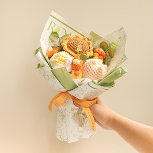 May include: A crocheted bouquet of sunflowers and peach-colored flowers wrapped in green and white patterned paper with a yellow ribbon.