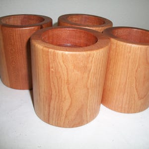 May include: A set of four cylindrical wooden cups with a smooth, light brown finish and visible natural wood grain patterns. Each cup features a hollow center and a thick, sturdy wall construction, arranged in a cluster on a flat surface.