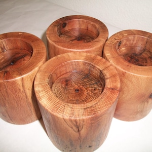 May include: Four handcrafted wooden candle holders. Each holder is cylindrical with a hollowed-out center to hold a candle. The wood has a warm, reddish-brown hue with visible grain patterns, showcasing the natural beauty of the material. The candle holders are approximately 3 inches tall.