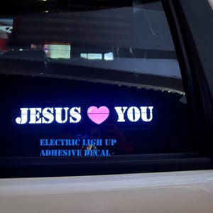 May include: An illuminated car window decal with the words "JESUS ❤ YOU" in white, with a pink heart symbol. Below, the text reads "ELECTRIC LIGHT UP ADHESIVE DECAL."