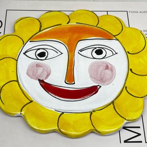 May include: A ceramic wall plaque shaped like a sun with a white face, a bright yellow scalloped border, and a central orange stripe. The face features black outlined eyes, pink circular cheeks, and a wide, curved red mouth painted on the surface.