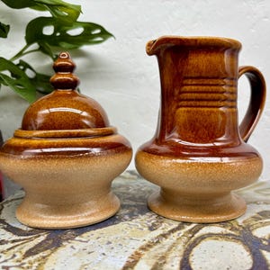 May include: A brown ceramic sugar bowl with a lid and a matching creamer jug. The sugar bowl has a rounded base and a domed lid. The creamer has a handle and a spout. Both pieces have a two-tone colour scheme.