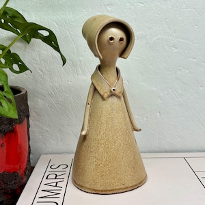 May include: A tan ceramic figurine of a person in a long dress and a bonnet. The figure has a simple design with two eye holes and a small button on the collar. The figurine is on a white surface.