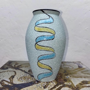 May include: A light blue ceramic vase with a textured surface and a black rim. The vase features a decorative design of intertwined yellow and blue ribbons outlined in black. A unique piece for home decor.