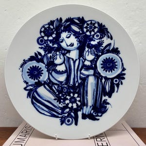 May include: White ceramic decorative plate with a blue illustration of a person surrounded by floral and geometric designs. The plate features a detailed, hand-painted style with a vintage aesthetic. The design is centered on the plate, creating a visually balanced composition.