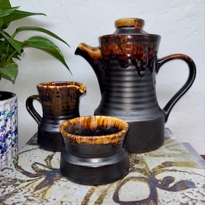 May include: A ceramic coffee set comprising a tall, dark brown coffee pot with a lid, a creamer, and a small bowl. The set has a textured, glazed finish with brown and tan accents. The pieces are arranged on a patterned surface.