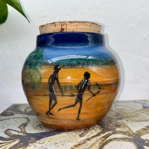 May include: A ceramic jar with a cork lid, depicting a painted landscape scene with two figures. The jar has a blue rim and a yellow and orange background, with a blue sky. The figures are black with white dot details.