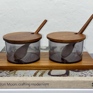 May include: Two glass spice jars with wooden lids and spoons on a wooden tray. The jars have a smoky grey tint and are filled with a light-coloured substance. The spoons and lids are made of a dark wood.