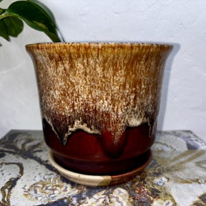 May include: A glazed ceramic plant pot in shades of brown and cream, with a deep red base and matching saucer. The pot features a flared rim and a textured, dripped glaze effect. A houseplant is visible in the background.