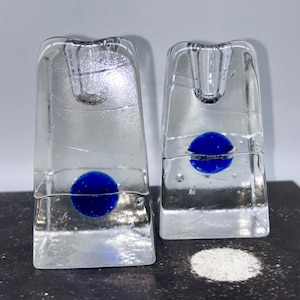 May include: Two clear glass candle holders, each with a blue orb. The candle holders are rectangular with a flared top and a small indentation for a candle. The glass has a textured appearance. The candle holders are made of glass.