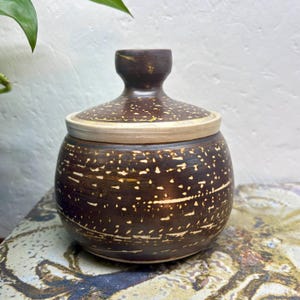 May include: A round, dark brown ceramic jar with a matching lid featuring a knob handle. The exterior surface is decorated with a repeating pattern of small, horizontal, light-coloured dashes and speckles arranged in horizontal bands around the body.