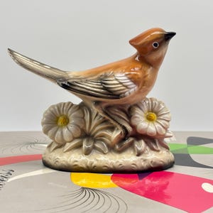 May include: A ceramic bird ornament with a crest, perched on a base with flowers. The bird has brown and tan feathers, a black beak, and a white eye. The flowers are white with yellow centres. The ornament is a decorative item.