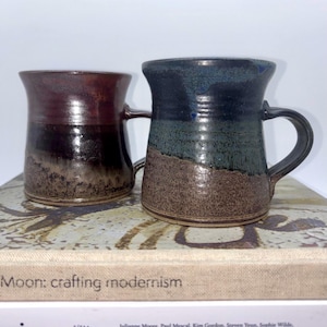 May include: Two handcrafted ceramic mugs with handles. One is a deep burgundy colour, the other is a blend of blue, green, and brown. The mugs are resting on a book titled "Moon: crafting modernism".
