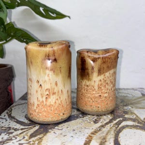 May include: Two handmade ceramic vases with a cylindrical form. The vases feature a distinctive glaze with a gradient of colours, including brown, tan, and orange. The texture appears slightly rough, enhancing the rustic aesthetic. The vases are displayed on a decorative surface.