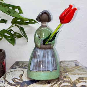 May include: A ceramic figurine of a woman in a green dress and apron, holding a red tulip. The figurine has a brown head and a grey hat. The tulip has green leaves and a long stem. The figurine is on a patterned surface.