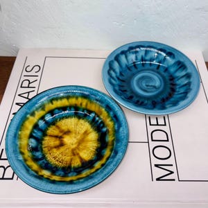 May include: Two ceramic bowls with a blue and yellow swirl design. The bowls have a glossy finish and a circular shape. The bowls are placed on a white surface with the word "MARIS" and "MODE" printed on it.