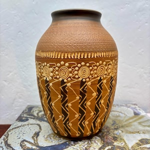May include: A brown ceramic vase with a wide, rounded body and a narrow neck. The surface features intricate, light-coloured etched patterns, including horizontal bands, circular spirals, and vertical zigzag lines over dark brown stripes.