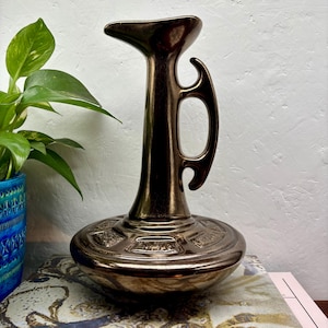 May include: A metallic bronze ceramic pitcher with a tall, slender neck, a flared spout, and a curved handle. The base is wide and circular, featuring a series of embossed rectangular panels with textured details around the lower section.