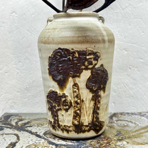 May include: A ceramic vase with a cream-coloured glaze and a textured, dark brown relief design depicting three stylised trees. The vessel features a rounded body, a narrow neck, and two small, integrated handles at the top rim of the piece.