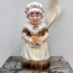 May include: A ceramic figurine of a woman in a chef's hat and apron, holding a bowl. The figurine is painted in shades of brown, white, and yellow. Wooden spoons are in the background. The figurine is on a decorative surface.