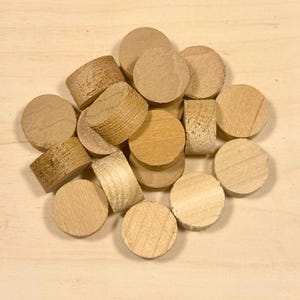 May include: A collection of unfinished wooden plugs and dowels in various sizes and shapes, scattered on a light-colored wooden square. The plugs are cylindrical and circular, with visible wood grain. The background is a wooden surface.
