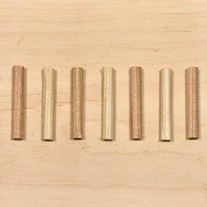 Op de afbeelding: Zeven houten deuvels in verschillende tinten lichtbruin en beige zijn horizontaal gerangschikt op een licht houten oppervlak. De deuvels zijn cilindrisch met een geribbelde textuur.