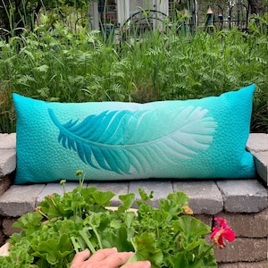 Könnte beinhalten: Ein türkisfarbenes und weißes gestepptes Lendenkissen mit einem großen weißen Federdesign. Das Kissen liegt auf einer Steinterrasse mit grünen Pflanzen im Vordergrund.