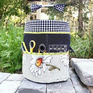 May include: A black and white checkered fabric bucket with a black and white polka dot fabric lining. The bucket has a white handle with black and white checkered bows on each end. The bucket is filled with sewing supplies, including scissors, a rotary cutter, and a ruler. The bucket is sitting on a stone surface.