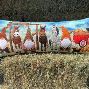 Pode incluir: Uma almofada longa e retangular com fundo azul e uma cena fantasiosa de gnomos, cavalos e uma carroça vermelha com abóboras. Os gnomos usam chapéus vermelho, branco e azul e têm barbas brancas. Os cavalos são castanhos, brancos e pretos. A carroça está cheia de abóboras e tem um gato preto no topo.